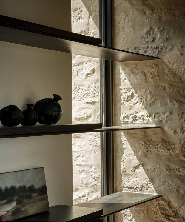 Close-up interior view of built-in open shelving beside a narrow floor-to-ceiling window. Dark timber shelves contrast with a rough cream stone masonry wall and pale plaster backing, displaying black sculptural vessels and a framed artwork. Strong natural light creates shadows across the textured stone.