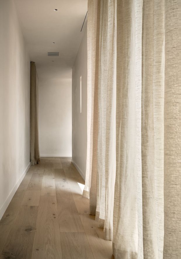 A narrow, minimalist hallway with pale timber plank flooring, smooth off-white walls and ceiling, and full-height sheer linen curtains along the right side. Soft natural light filters through the curtains, creating a calm, warm and understated interior. Recessed ceiling lights and a discreet linear air vent provide subtle functional detailing.