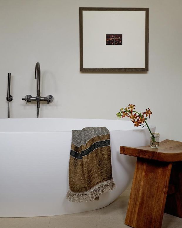 A minimalist bathroom featuring a freestanding white bathtub with a wall-mounted chrome bath and shower fixture. A textured brown, grey, and black towel hangs over the tub, while a sculptural timber side table with a small vase sits beside it. A framed artwork adds a subtle decorative element to the pale wall, and large light neutral floor tiles complete the room.