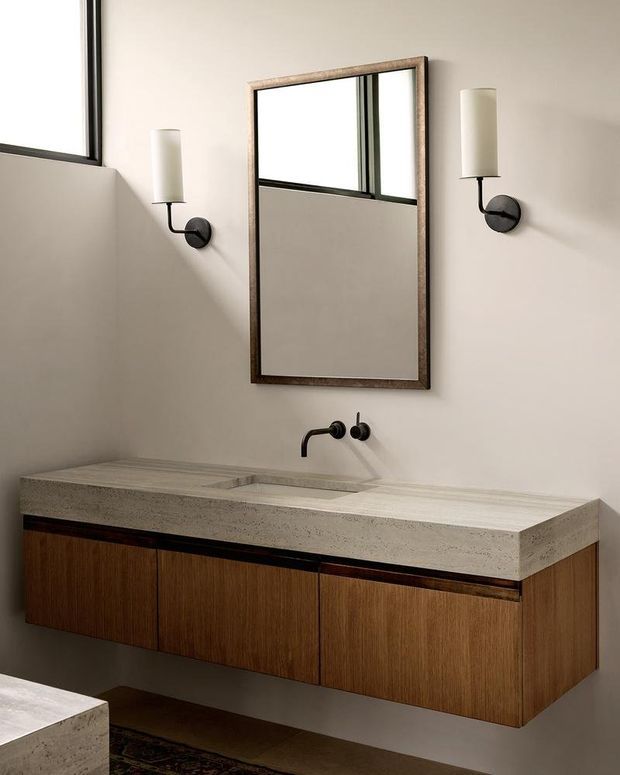 A refined contemporary bathroom featuring a floating timber vanity with warm vertical-grain cabinetry, a thick light stone integrated basin benchtop, matte black wall-mounted tapware, and matching black-and-white wall sconces. A tall aged-metal framed mirror sits above the vanity, with a high window providing natural light. The room has a warm neutral palette and clean, minimalist detailing.