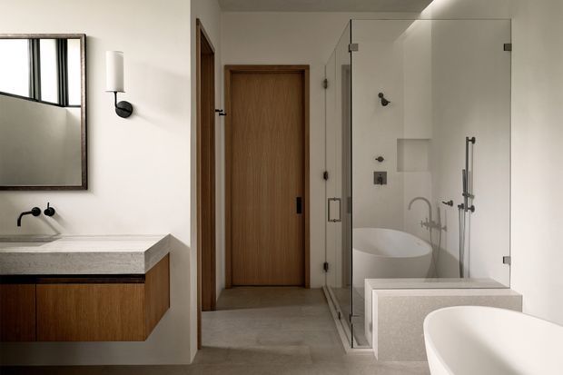 Contemporary bathroom with a freestanding bathtub in the foreground, a glass-enclosed shower and second tub area, and a wall-mounted timber vanity with a pale stone benchtop. The room has white walls, concrete-look flooring, black plumbing and lighting fixtures, a timber door, and a large mirror.