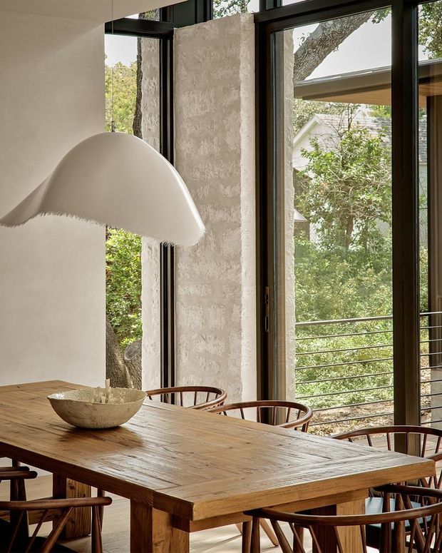 Warm, refined dining area with a large rustic timber dining table and curved wooden chairs beside expansive black-framed glazing. Textured pale plaster columns and walls contrast with the natural timber, while leafy trees and an adjacent structure are visible outside. A sculptural white pendant light hangs over the table, creating a calm indoor-outdoor connection.
