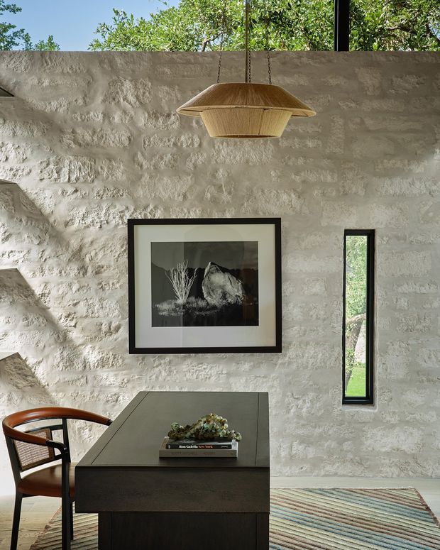 A refined interior vignette featuring a dark built-in or freestanding desk beneath a woven pendant light. The room has a heavily textured pale stone or rendered masonry wall, narrow black-framed vertical window, framed monochrome artwork, a timber-and-leather chair, and a striped area rug. Lush greenery is visible through the high glazing and window, creating a warm connection to the outdoors.