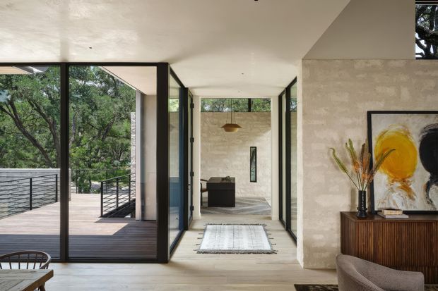 A bright contemporary interior opening onto a timber deck through large black-framed glazed doors. The space features pale timber flooring, textured light walls, a narrow corridor leading to a sitting area, a fluted dark-wood sideboard, abstract artwork, and restrained natural decor. A leafy landscape is visible outside.