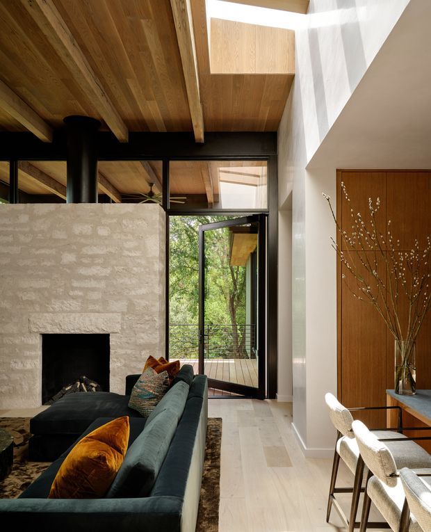 A warm, contemporary open-plan living and dining space with a dark green sectional sofa, textured stone fireplace, timber-lined ceiling, black-framed glazing, and a timber pivot door. Full-height glazing and an open door connect the room to a leafy exterior deck, while a compact dining area sits beside a white plaster wall. Natural timber, stone, and strong sunlight create a refined indoor-outdoor atmosphere.