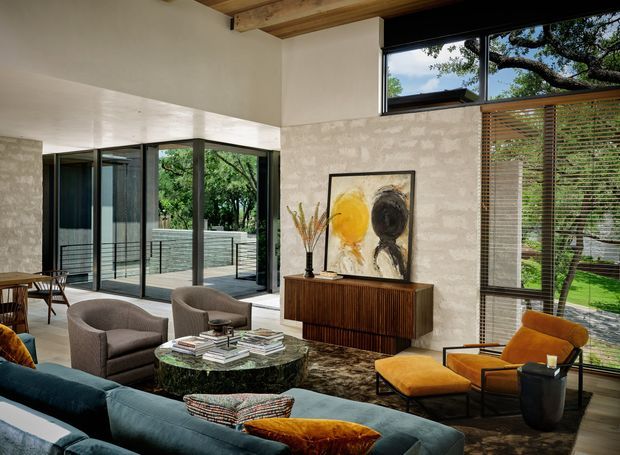 A spacious, light-filled living room with floor-to-ceiling black-framed glazing opening to a landscaped terrace, exposed timber ceiling beams, pale textured walls, and timber flooring. The room features a deep blue sectional sofa, upholstered lounge chairs, a sculptural dark stone coffee table, a ribbed timber sideboard, artwork, and warm mustard accent seating. Horizontal timber blinds and abundant greenery create a strong indoor–outdoor connection.