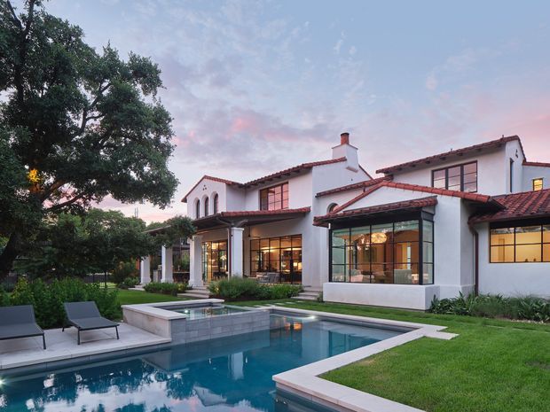 Exterior view of a large two-storey white stucco residence with red clay-tile pitched roofs, arched openings, dark-framed glazing, and a glazed sunroom-like extension. The landscaped backyard features a rectangular swimming pool, paved pool surrounds, lawn, mature trees, and lounge furniture, photographed at dusk.
