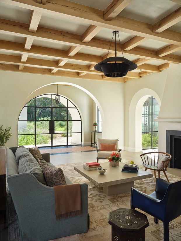 A refined living room with cream plaster walls, large arched black-framed windows and doors, a substantial fireplace, and a layered seating arrangement around a square stone coffee table. The ceiling features prominent exposed timber beams arranged in a coffered grid, complemented by a dramatic dark pendant light. Neutral upholstery, patterned textiles, and a geometric rug create a warm, elegant interior.