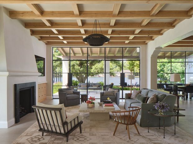 A spacious, refined living room with a large fireplace, exposed timber coffered ceiling, expansive black-framed glazing, and an open connection to a dining area. The room is furnished with a muted green sofa, mid-century-style lounge chairs, a stone or concrete coffee table, and a patterned neutral rug. Lush garden views are visible through the glazing.