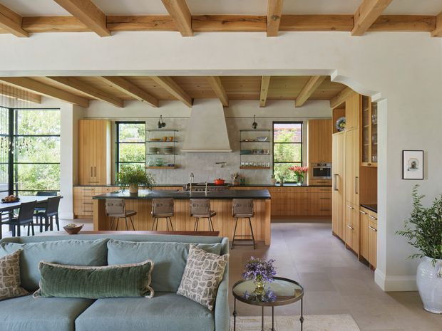 Open-plan kitchen and living area with a large timber island, integrated timber cabinetry, exposed natural timber ceiling beams, pendant lighting, and a pale blue upholstered sofa in the foreground. The kitchen includes a sculptural plaster rangehood, shelving, stainless appliances, and dark benchtops, while a dining area is visible to the left. The room has a warm, refined, natural aesthetic with abundant daylight.