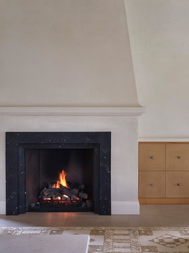 A refined interior detail centered on a traditional fireplace with a lit fire, framed in dark veined stone and set within a pale plaster mantel surround. Built-in light timber cabinetry with small dark knobs is visible to the right, while a neutral patterned rug and pale furnishings occupy the foreground. The palette is soft and warm with strong contrast from the black fireplace surround.