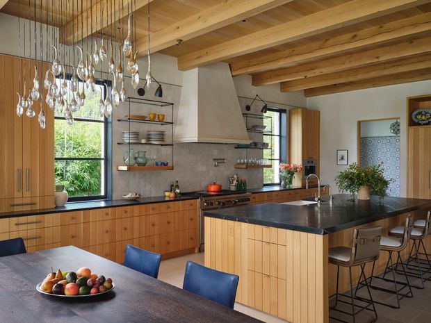 Warm, spacious open-plan kitchen and dining area with extensive light timber joinery, exposed timber ceiling beams, a large dark stone island, and a sculptural plaster rangehood. Black-framed windows, open shelving, pendant glass lighting, and blue dining chairs add contrast. The kitchen opens visually to a dining space and leafy outdoor view.