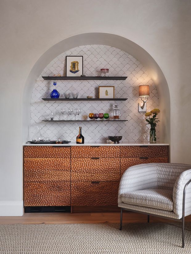A refined interior vignette featuring a recessed arched alcove with patterned tiled backing and three dark floating shelves displaying glassware, framed art and decorative objects. Below is a large copper-toned textured sideboard with black pulls and a pale stone or composite countertop. A sculptural wall sconce, vase of flowers and upholstered patterned armchair complete the warmly lit setting. Timber flooring and a neutral woven rug add warmth and texture.