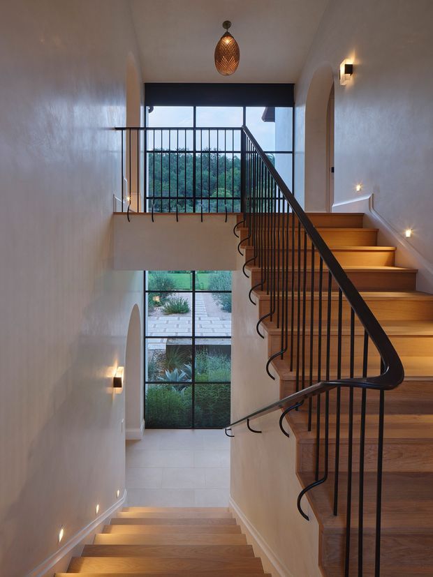 A contemporary multi-level stairway with warm timber treads, slim black metal balustrades, white textured walls, and sculptural wall and pendant lighting. Large black-framed windows bring in greenery and daylight, while an arched doorway adds a soft architectural detail.