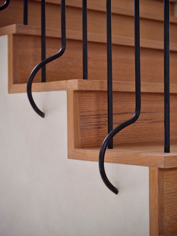 Close-up view of a timber staircase with warm wood treads, risers, and side stringers, set against a light painted wall. Slim black metal balusters run vertically along the stair edge, with curved decorative ends creating a strong graphic detail. The image focuses on stair construction and railing craftsmanship rather than a full room.