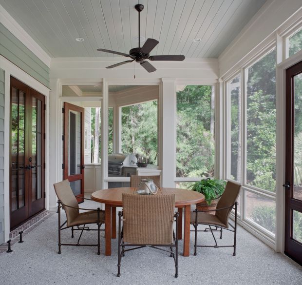 A bright enclosed dining and sunroom with a round wooden dining table surrounded by woven and upholstered chairs. The room features expansive screened or glazed windows overlooking greenery, dark-framed glazed doors, pale green and white wall panelling, a white beadboard ceiling, a dark wood ceiling fan, and a light speckled floor. An outdoor grill or kitchen area is visible through the rear opening.