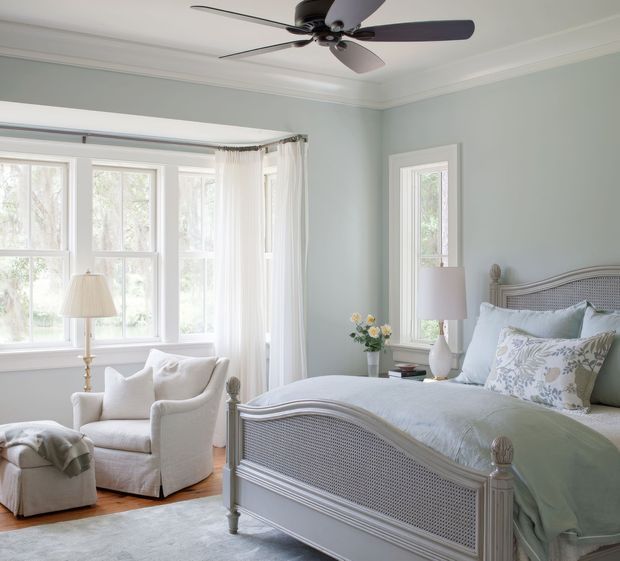 Bright, airy bedroom with a large upholstered and cane-panel bed, pale blue-grey walls, white trim, layered light bedding, and floral accent pillows. Tall windows with sheer curtains provide abundant natural light, while an armchair, ottoman, bedside tables, lamps, and a ceiling fan complete the calm traditional coastal-style interior.