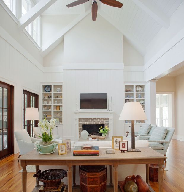 Bright, traditional-style living room with a dramatic vaulted white-painted ceiling, exposed white beams, and a large central fireplace wall with built-in display shelving and a television. Pale upholstered seating faces the fireplace, while a substantial timber console table with lamps, flowers, framed photographs, and books occupies the foreground. Tall glazed doors and windows provide abundant natural light, and a large dark timber ceiling fan hangs overhead.