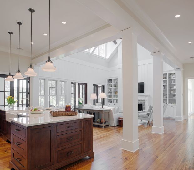 Bright open-plan interior combining a kitchen, living area and dining zone. The kitchen features a large dark timber island with a pale stone benchtop, pendant lights and stainless-steel tapware. White wall panelling, built-in shelving, substantial white columns, timber flooring and a skylight create an airy, traditional-inspired open living space.