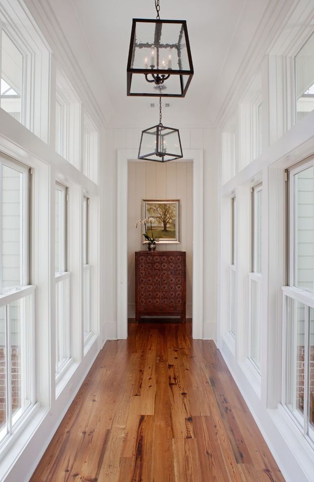 A bright, narrow residential hallway or gallery corridor with extensive white-painted trim and wainscoting, tall multi-pane windows on both sides, and warm timber flooring. Two black metal and glass lantern pendant lights hang along the central axis. At the far end, a dark carved wooden console cabinet supports an orchid beneath a framed landscape artwork. The space has a refined traditional character with strong symmetry and abundant natural light.