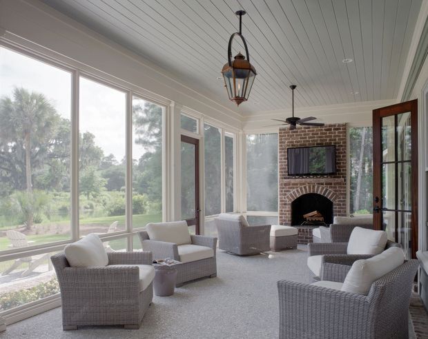 Bright enclosed sunroom with extensive floor-to-ceiling glazing, a white beadboard ceiling, pale carpet, and multiple light woven lounge chairs arranged around a central coffee table. A red brick fireplace with arched detailing and mounted television forms the far feature wall, while lantern pendant lights and a ceiling fan add a relaxed traditional coastal character. Views of greenery are visible through the windows.