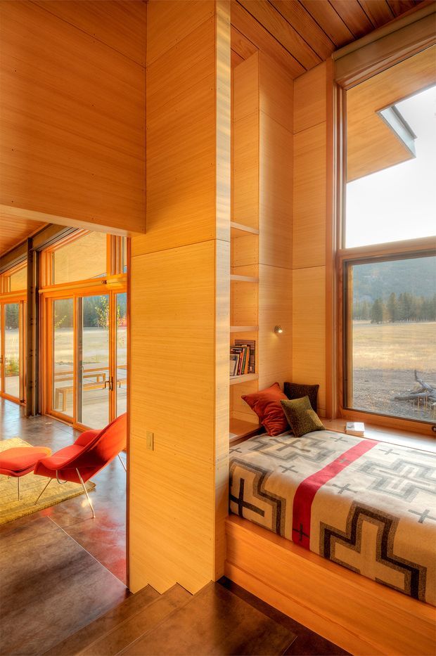Warm, wood-lined interior featuring a built-in window seat with patterned blanket and cushions, integrated shelving, expansive timber-framed glazing, and a small sitting area with bright red lounge chairs. The room opens toward a glazed exterior connection and overlooks a natural rural landscape. Rich timber surfaces, polished concrete flooring, and strong natural light create a cozy contemporary retreat.