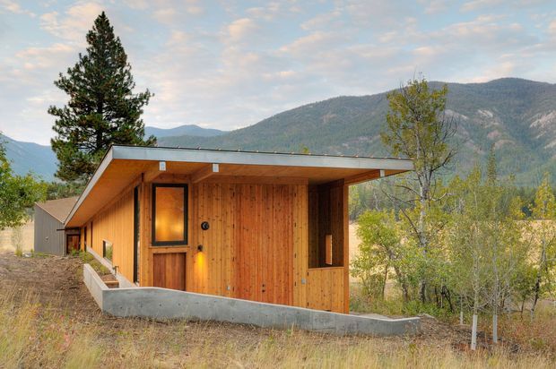 A compact single-storey timber-clad cabin set on a sloping site, with a broad low-pitched roof, recessed porch, and simple dark-framed glazing. The building is surrounded by dry grass and trees, with a mountainous landscape visible behind it. The exterior composition emphasizes warm vertical timber boards, concrete edging, and a minimalist lodge-like form.