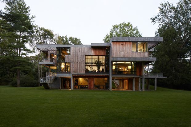 Exterior view of a contemporary multi-level home set within a wooded landscape. The building has a strongly horizontal, rectilinear composition with projecting upper-floor volumes, extensive glazing, open terraces and balconies, and warm vertical timber cladding. The surrounding lawn and mature trees frame the house, while illuminated interiors are visible through the windows.