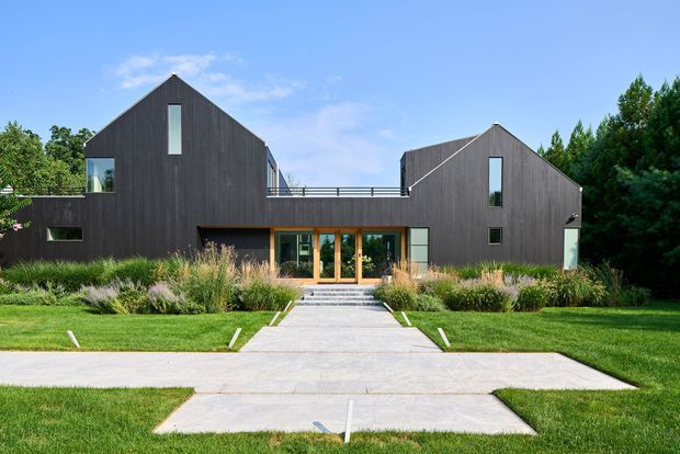 Front exterior view of a contemporary multi-volume house with two steep gabled wings clad in dark vertical boards. The central recessed entry has warm timber-framed glazing and opens onto a broad pale paved walkway, with clipped lawns and ornamental grasses forming a minimalist landscaped approach.