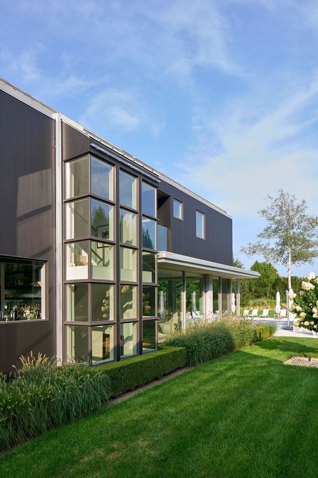 Exterior view of a contemporary two-storey residence with dark vertical cladding, extensive floor-to-ceiling glazed corner windows, a projecting covered terrace, and a carefully landscaped lawn with clipped hedges and ornamental grasses. A glimpse of an interior bar is visible through the glazing.