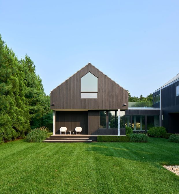 A modern two-storey residential exterior set within a landscaped garden. The central gabled volume is clad in dark vertical timber, with a large geometric upper window and a covered ground-floor terrace furnished with two chairs. A glazed connecting section links it to a darker flat-roofed wing on the right. The foreground is an expansive manicured lawn, with dense screening vegetation to the left and low ornamental planting around the building.
