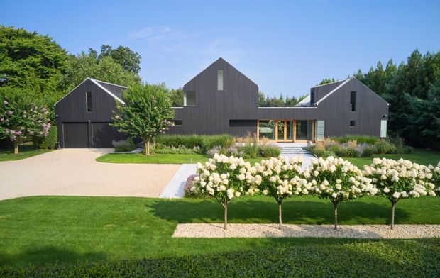 A contemporary multi-wing residential home viewed from the landscaped front garden. The building has dark vertical cladding, strongly geometric gabled forms, narrow vertical windows, and a glazed central living area. A broad driveway leads to an integrated garage, with manicured lawns, clipped flowering shrubs, and mature trees surrounding the property.