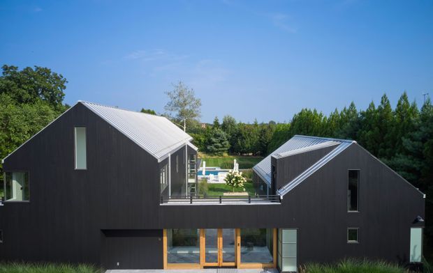 Exterior view of a contemporary dark-clad residence composed of two pitched-roof volumes connected by a recessed central terrace or bridge. Full-height glazing and warm timber-framed entry doors open toward a landscaped garden and pool visible beyond. The building has crisp geometric forms, charcoal vertical cladding, pale metal roofing, and a sheltered courtyard-like connection between the wings.