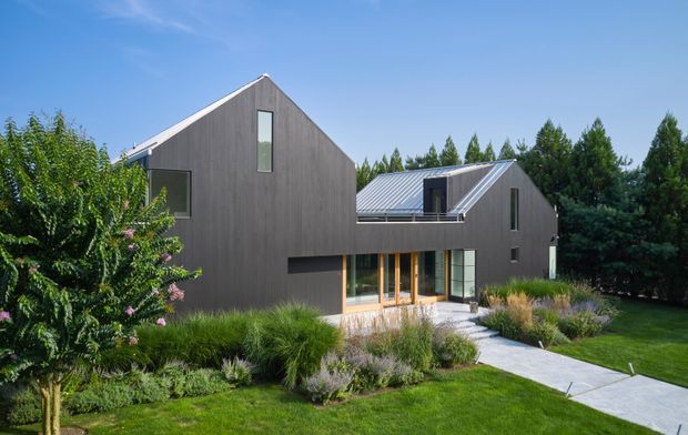 Exterior view of a contemporary multi-volume house with dark vertical timber cladding, steep gabled roofs, and extensive glazing. The building is set within a landscaped garden with ornamental grasses, shrubs, paved paths, and mature trees under a clear blue sky.