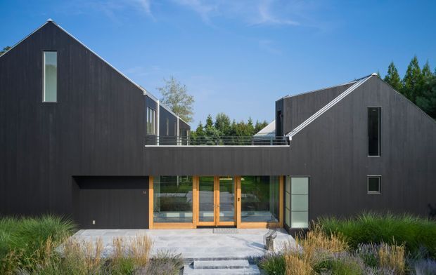 A contemporary dark-clad residential building with two steeply pitched gabled volumes connected by an elevated central bridge or terrace. The facade features slim vertical windows, warm timber-framed full-height glazing at the ground-floor entry, and a pale paved approach surrounded by ornamental grasses and low landscaping under a clear blue sky.
