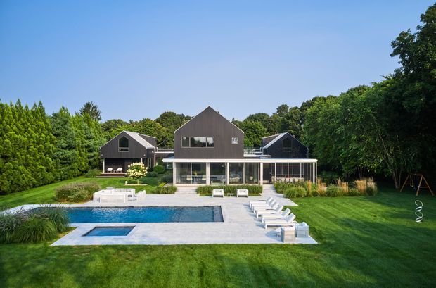 A contemporary multi-wing residential house viewed from the landscaped rear garden, with dark grey gabled volumes, extensive ground-floor glazing, a large paved terrace, and a rectangular swimming pool with spa in the foreground. Mature trees and clipped lawn surround the property.