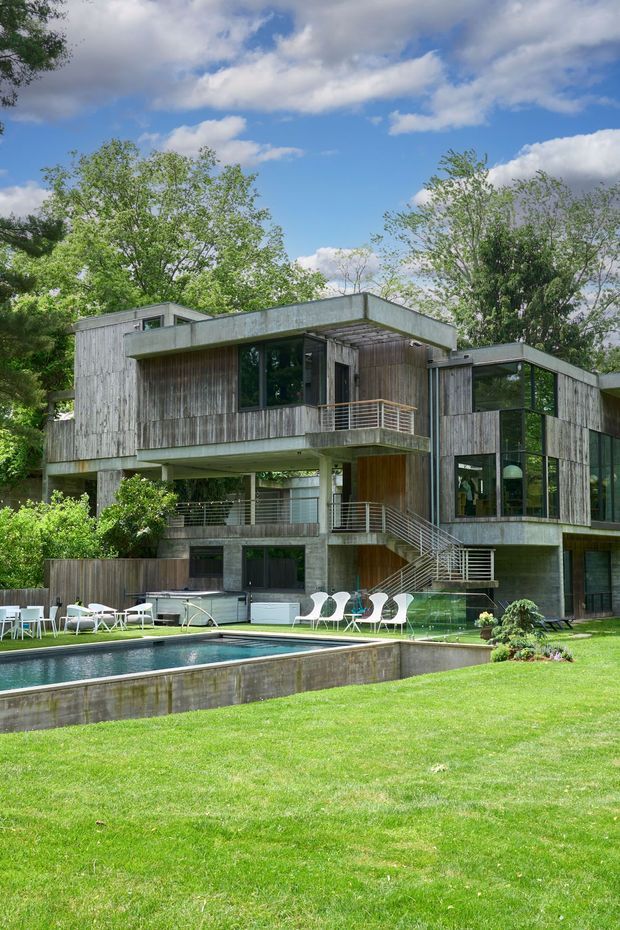 Exterior view of a modern multi-level residence set within a leafy landscaped garden. The house features flat rooflines, projecting and cantilevered volumes, extensive glazing, weathered vertical timber cladding, exposed concrete elements, and open metal balustrades. A rectangular swimming pool and outdoor dining area occupy the foreground.