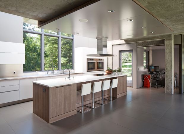 Contemporary open-plan kitchen with a large wood-finished island, white waterfall-style benchtop, integrated appliances, and four white bar stools. Floor-to-ceiling glazing brings in views of greenery, while a concrete structural column, exposed concrete ceiling, dark trim, and large grey floor tiles create a refined modern-industrial character. A home office or utility space is visible beyond the kitchen.