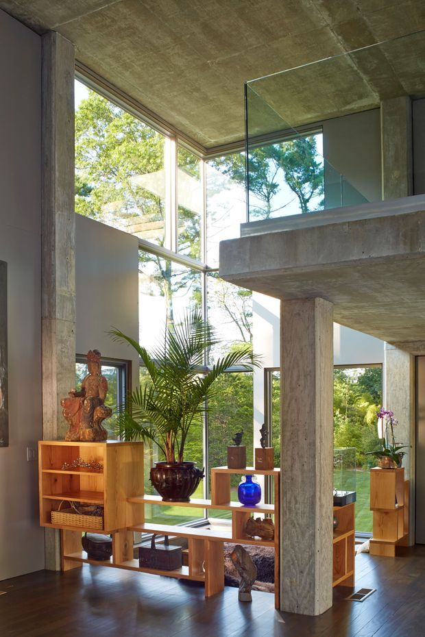 A double-height modern interior with exposed raw concrete ceilings, columns and beams, extensive floor-to-ceiling glazing, and a mezzanine-like upper level with a clear glass balustrade. Warm timber open shelving and cabinetry display decorative objects and plants, while dark timber flooring contrasts with the concrete structure. Lush greenery is visible through the windows.