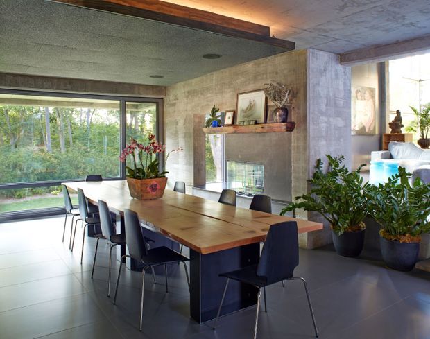 A contemporary dining room with a long timber dining table and dark upholstered chairs, positioned beside expansive windows overlooking wooded greenery. The room features concrete-look walls, a textured ceiling, a fireplace with a simple timber mantel, large potted plants, and a partial view into an adjacent living area.