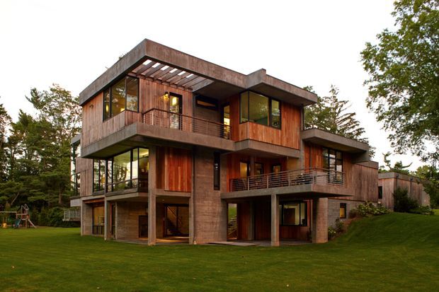 A multi-storey contemporary residential house set within a landscaped garden, featuring strong horizontal concrete forms, extensive glazing, warm timber cladding, open balconies, and a rooftop pergola-like projection. The building has an irregular stepped massing and appears to be integrated into a wooded suburban or rural setting.