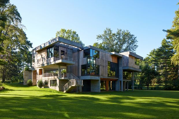 A contemporary multi-storey residence set within a spacious landscaped lawn and mature trees. The house has a strongly horizontal composition with flat roof planes, projecting balconies, extensive dark-framed glazing, exposed concrete elements, and vertical timber cladding. Exterior stairs and elevated sections reinforce the modern structural character.