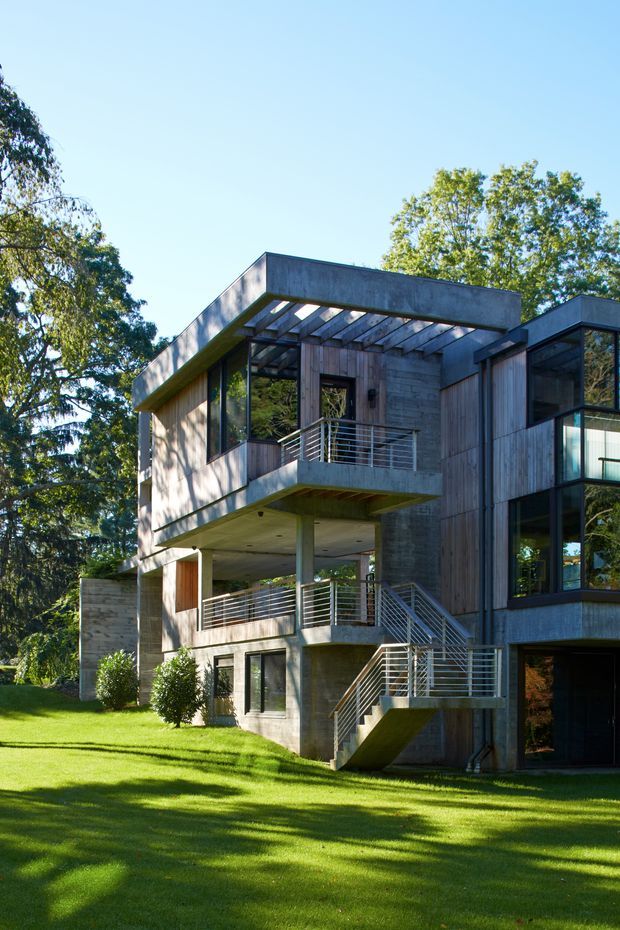 Exterior view of a contemporary multi-level house set in a landscaped, tree-lined site. The building features exposed concrete walls and slabs, extensive floor-to-ceiling glazing, vertical timber cladding, projecting box forms, open pergola-like roof framing, and exterior metal staircases and balustrades.