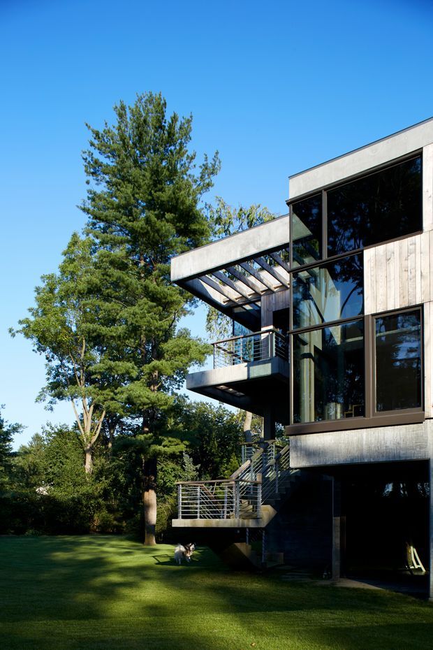 Exterior view of a contemporary multi-level house set within a mature landscaped garden. The building features strong rectangular volumes, extensive dark-framed glazing, pale vertical timber cladding, projecting concrete-edged balconies, open metal balustrades, and a partially covered terrace or pergola structure. Bright blue sky and established trees surround the home.