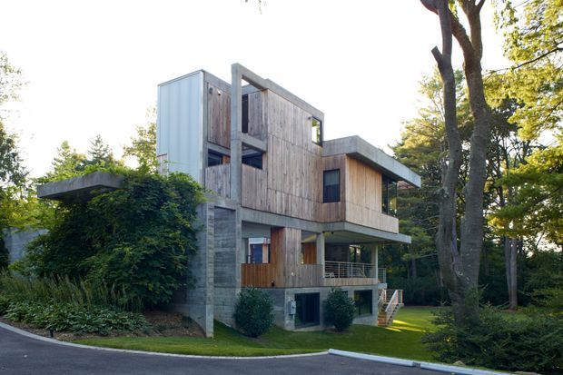 A modern multi-level residence set among mature trees, featuring a strongly articulated rectangular form, exposed concrete structural walls, extensive vertical timber cladding, dark-framed glazing, projecting balconies, and flat roof planes. The house is viewed externally from a landscaped suburban site.