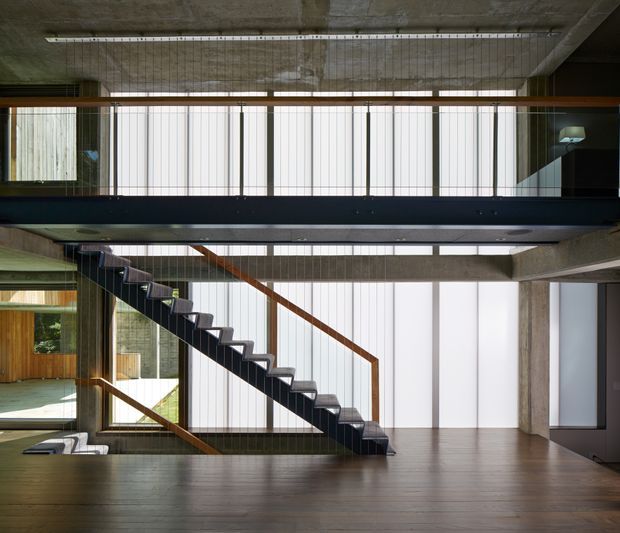 Interior view of a modern multi-level home focused on a dramatic open stairway. A dark steel stair with timber handrails crosses in front of tall translucent vertical glazing, connecting a lower timber-floored level with an upper mezzanine or landing. Exposed concrete ceilings and beams, dark metal balustrades, and extensive daylight create an industrial yet refined architectural character.