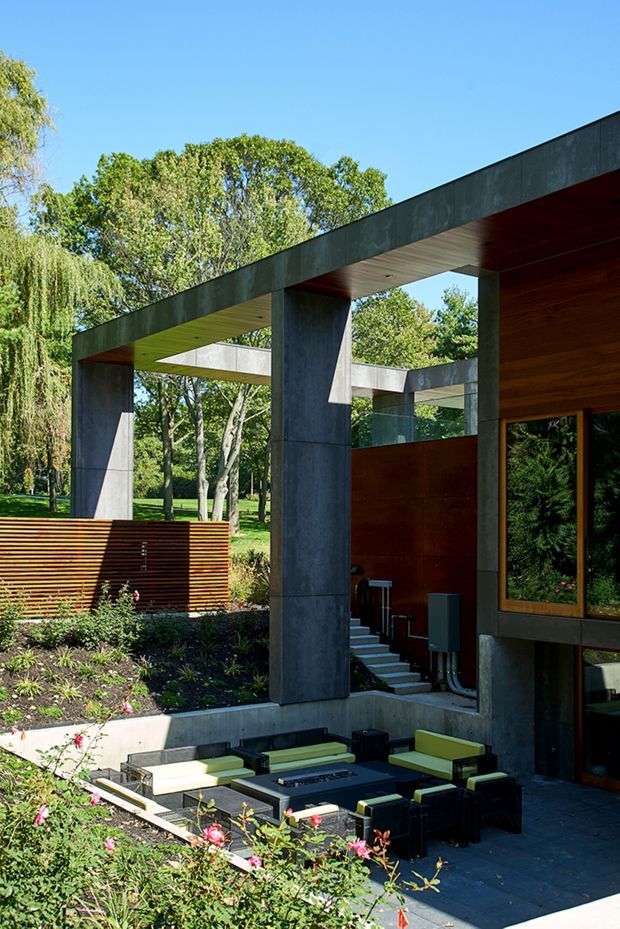 Exterior architectural view of a modern residence set within a landscaped garden. The composition features strong concrete columns and beams forming open flat-roofed terraces, warm timber wall cladding and screening, large dark-framed windows, and a sunken outdoor lounge with low seating around a central fire or coffee table. Mature trees and planting surround the building.