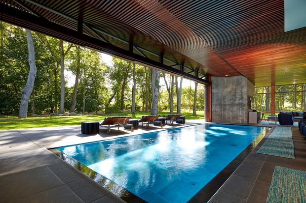 A sophisticated indoor-outdoor pool pavilion with a rectangular swimming pool extending beneath a dramatic open-sided roof. The space features floor-to-ceiling glazing, slender dark structural framing, a richly lined timber slatted ceiling, concrete walls, large dark stone or concrete floor tiles, and lounge chairs arranged around the pool. The pavilion opens directly onto a landscaped wooded setting, creating a bright, resort-like atmosphere.