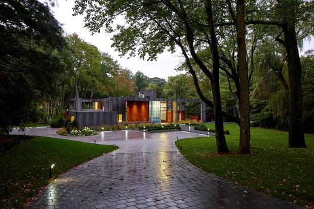 A low, contemporary single-storey residence viewed from a wet paved driveway through a mature, heavily landscaped garden. The home has a strongly horizontal composition with dark vertical cladding, extensive glazed sections, warm illuminated interior spaces, and a flat or very low-pitched roofline. Large established deciduous and weeping trees frame the approach, with curved planting beds and discreet path lighting.