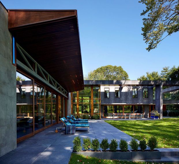 A contemporary courtyard-style residence with strong horizontal volumes, extensive floor-to-ceiling glazing, dark metal or timber cladding, and broad overhanging roof planes. The paved terrace includes outdoor lounge seating and raised planter beds, while the central lawn and mature trees create a landscaped outdoor setting. Exposed structural trusses and warm timber-framed glazing add architectural detail.